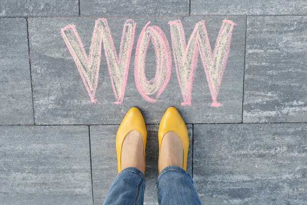 Wow written on gray sidewalk with woman legs in feet in yellow shoes