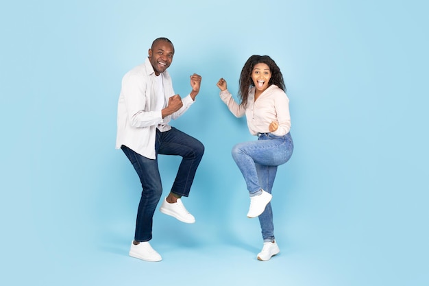 Wow yes Emotional black man and woman shaking clenched fists looking at camera with excitement blue studio wall