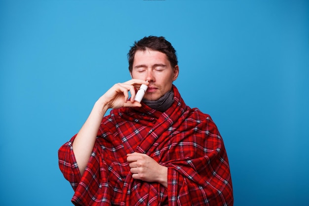 A wrapped in a blanket sick man is standing and using nasal spray