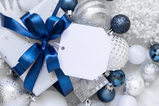 Wrapped Gift Box with a blue bow and paper gift tag on a white table with white and silver Christmas decorations around top view. Winter composition with blank label card, Mockup, copy space