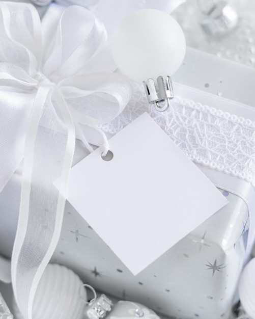 Wrapped present with a white bow and square paper gift tag on a white table with white and silver Christmas decorations close up. Winter composition with blank label card, Mockup, copy space