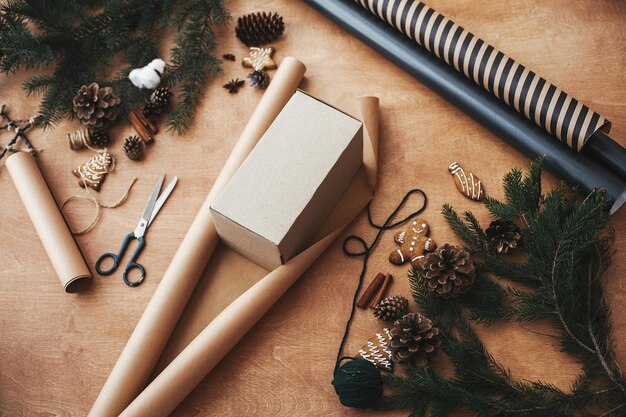 Wrapping christmas gifts concept empty box on stylish craft\
paper and pine branches cones gingerbread cookiesthread cinnamon\
scissors on rural wooden table seasons greeting