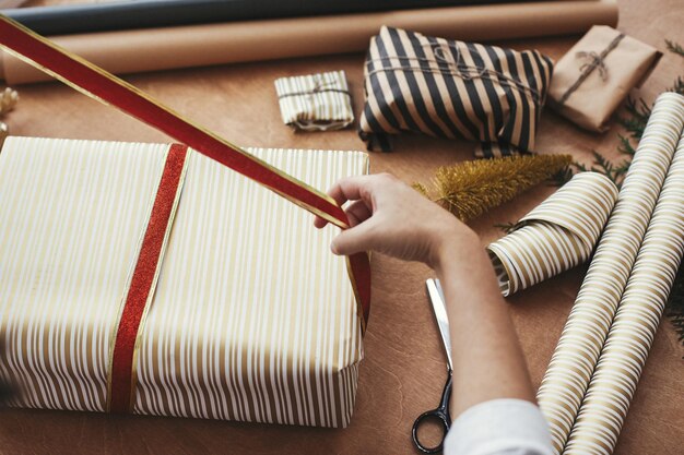 Wrapping christmas gifts concept hands holding red ribbon and\
wrapping stylish gift box in striped golden paper and scissors\
presents golden tree on wooden table