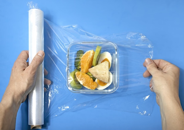 Wrapping with a plastic film a tapperware with takeaway food on\
a blue background