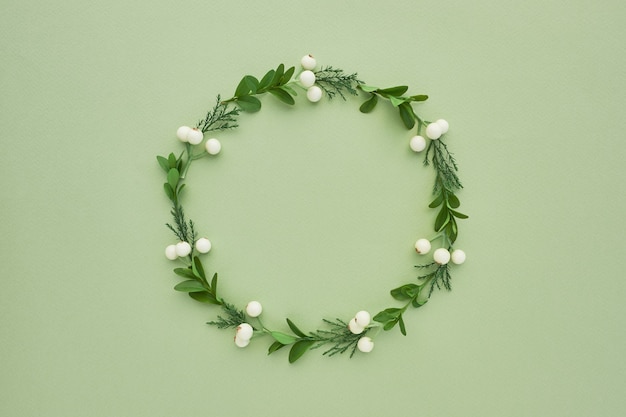 Wreath frame made of evergreen tree branches and white berries on green background. Top view, flat lay.