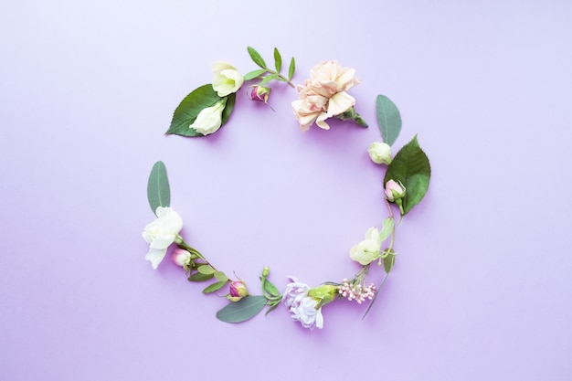 A wreath in a round frame with rosebuds, delicate white flowers, branches, leaves and petals on a purple background. Flat layout, top view