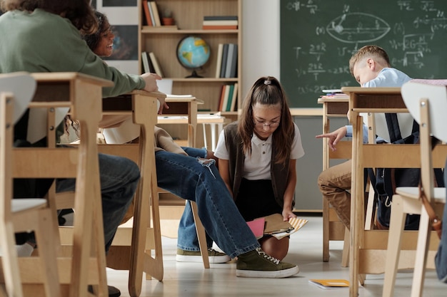 Wrede schoolkinderen lachen om klasgenoot die op knieën in gangpad tussen bureaus staat