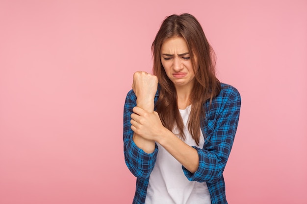 Wrist pain. portrait of unhappy sick girl in checkered shirt\
holding her injured hand and grimacing from pain, carpal tunnel\
syndrome, pinched nerve. indoor studio shot isolated on pink\
background