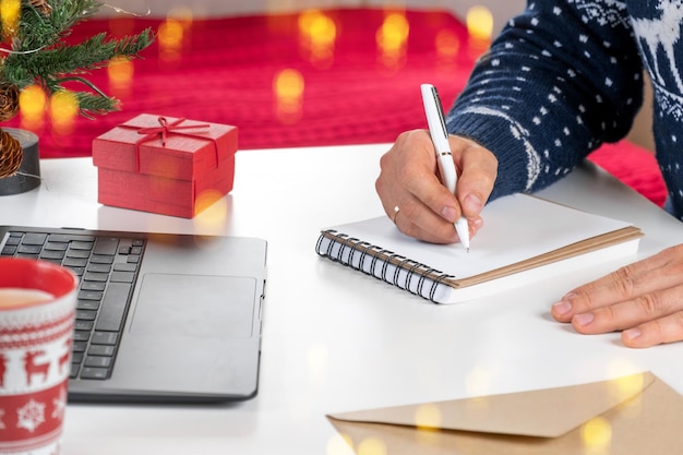 Foto un biglietto di auguri di natale maschio adulto che scrive carte di auguri in ufficio a casa durante il natale uomo mani in maglione blu che tiene carta bianca e busta alla decorazione natalizia della tazza di caffè desktop