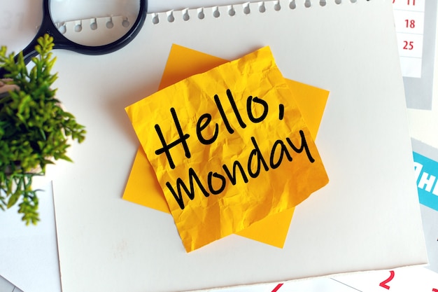 Writing on crumpled yellow paper: Hello Monday. Defocus. Black letters. Magnifier and green potted plant on the desk office.