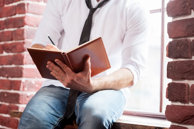 Writing down his thoughts. Close-up of young man in shirt and tie writing something in note pad while sitting at the window sill