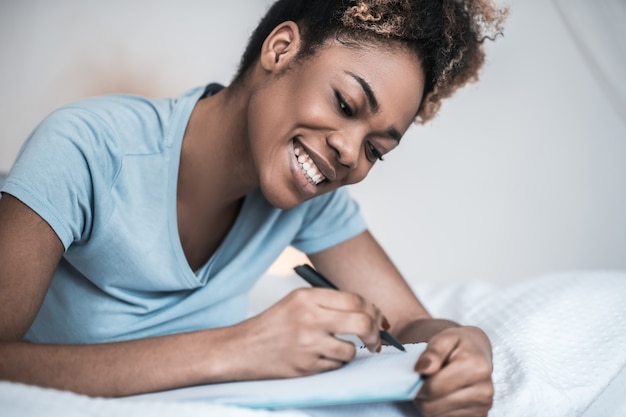 Writing thoughts. Smiling young adult african american woman writing with pen in notebook lying on stomach on bed