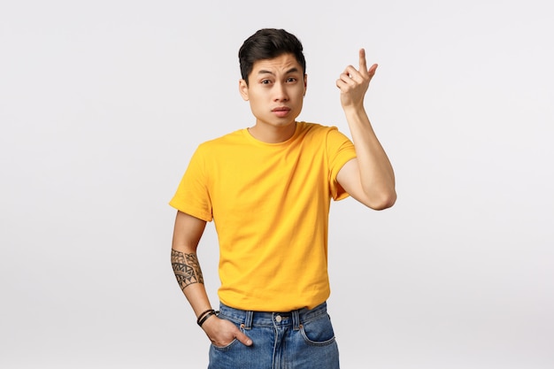 Wtf aan de gang. Geërgerd Aziatische jongeman in geel t-shirt, met tatoeages, wijsvinger in wanhoop en irritatie opheffen, wenkbrauwen optrekken als onzin horen, raar gesprek, witte muur
