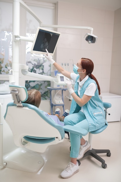 Рентген на экране. Рыжий детский стоматолог в маске и униформе показывает девушке рентгеновский снимок на экране