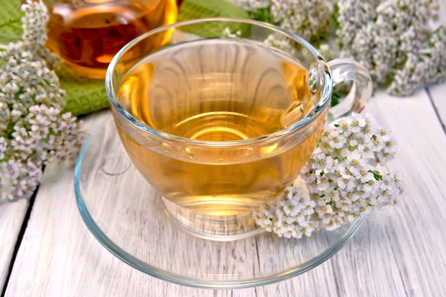 Tè di achillea in una tazza di vetro e teiera, fiori freschi di achillea su uno sfondo di tavola di legno chiaro