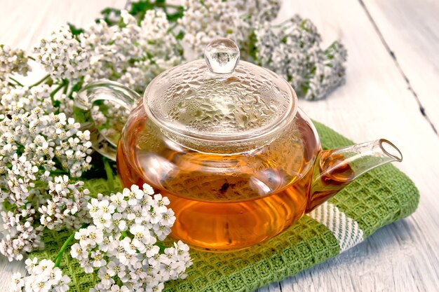 Yarrow tea in a glass teapot on a green napkin, fresh yarrow flowers on a of pale wooden plank