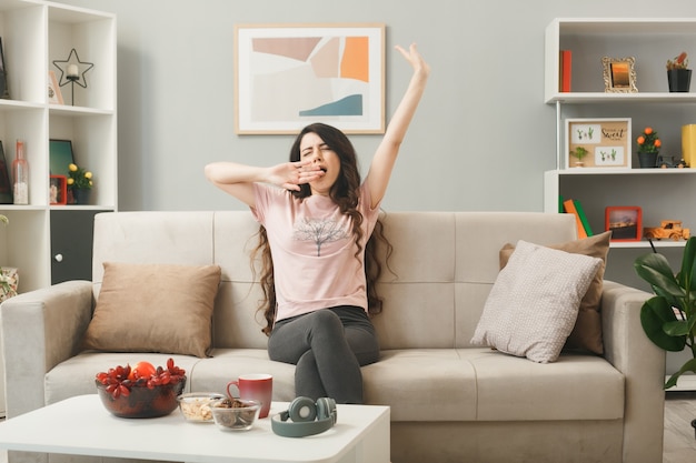 Yawning covered mouth with hand young girl sitting on sofa behind coffee table in living room