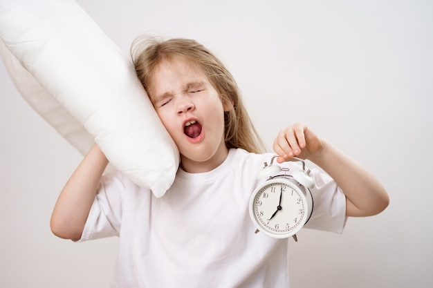 A yawning girl hugs a pillow and an alarm clock on white