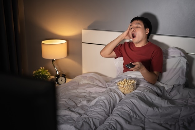 Yawning sleepy man man watching TV movie on a bed at night