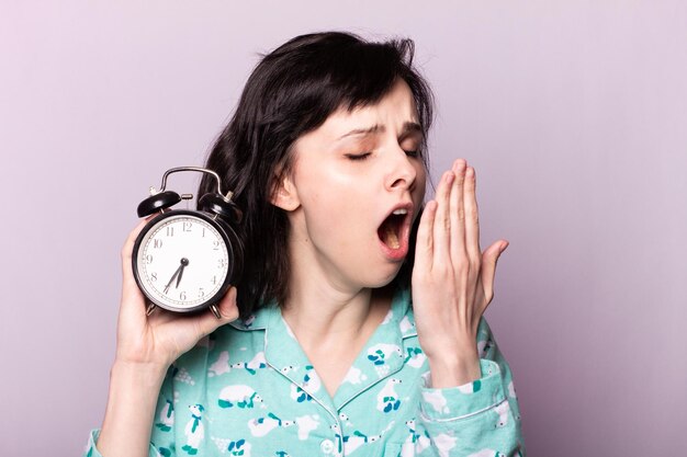 パジャマのあくびをする女性は手に目覚まし時計を持っています