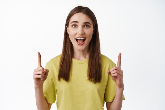 Yeah big news comming ahead. Excited cute girl scream from joy and amazement, pointing fingers up, making announcement, showing discount banner, white background