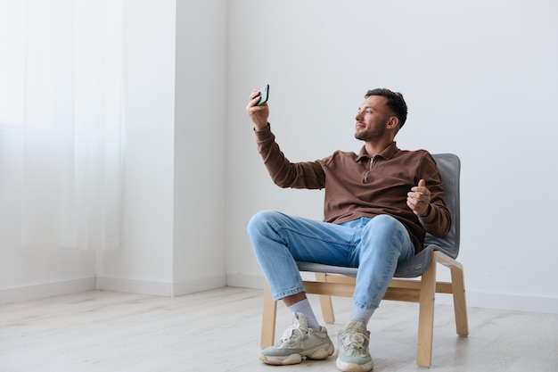 Yeah cool offer smiling cheerful overjoyed happy young tanned
man doing selfie video call hold phone sitting on chair at home
near white wall distance communication concept copy space
