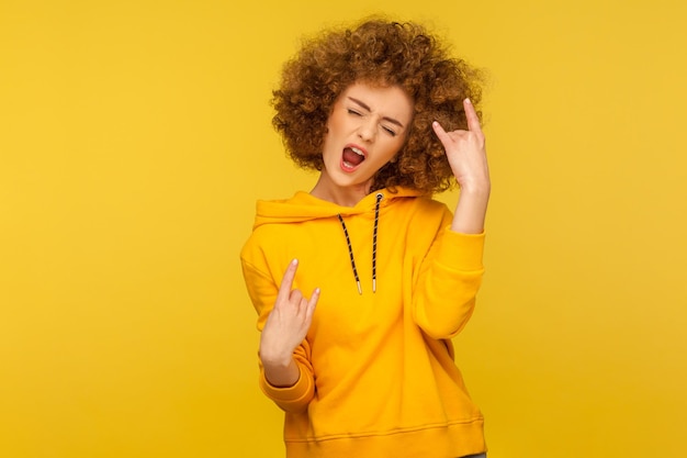 Yeah, thats wonderful. Portrait of overjoyed curly-haired woman in urban style hoodie showing rock and roll hand sign, screaming and gesturing to heavy metal, rock music. indoor studio shot isolated