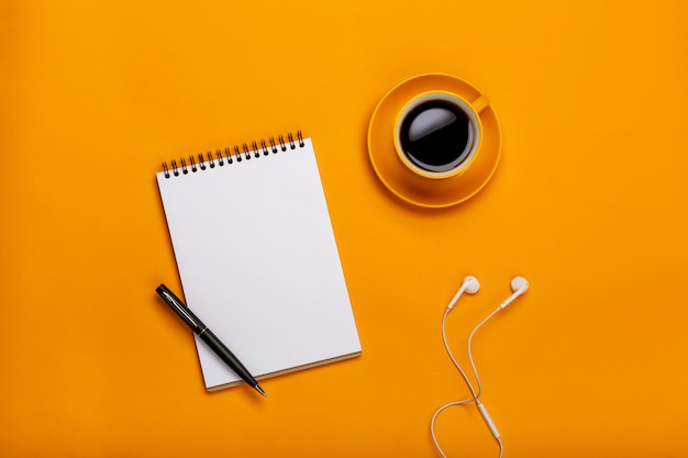 On a yellow background, a cup of black coffee with a notepad and headphones