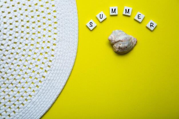 On a yellow background there is a shell and the word summer is written above it a summer hat is lyin...