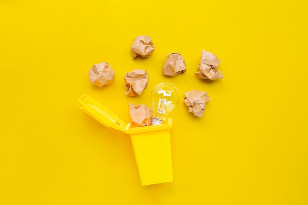 Yellow bin with light bulb and brown crumpled paper on yellow background. Ideas and creative thinking concept.