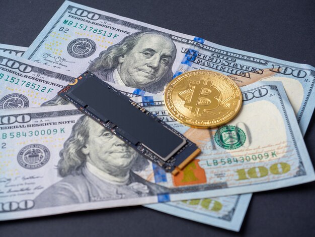 Yellow bitcoin, an m2 SSD disk and hundred-dollar bills lie on a black background. The concept of mining cryptocurrency through hard drives