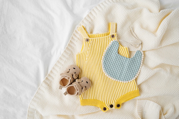 Yellow bodysuit, bib, baby boots and toy on knitted blanket. Set of  kids clothes and accessories  on bed. Fashion newborn. Flat lay, top view