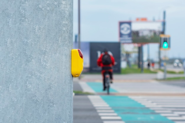 Pulsante giallo in corrispondenza di un semaforo per pedoni sullo sfondo di un passaggio pedonale e di un ciclista su una pista ciclabile.