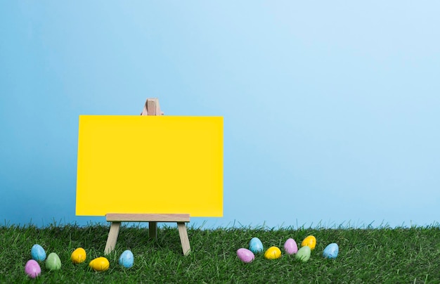 Yellow canvas on an easel place for text on the grass among easter eggs on the blue sky background