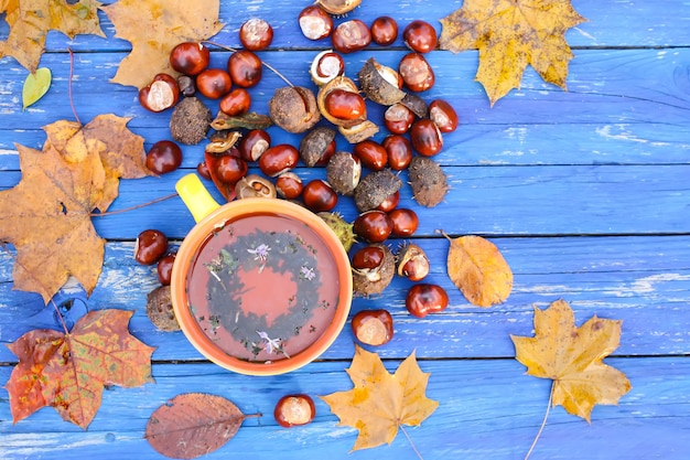 Yellow ceramic cup of herbal tea on aged wooden background with fall autumn leaves and chestnuts.