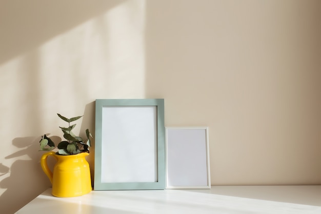 A yellow ceramic jug or vase with eucalyptus branches, empty white photo frames on the white table in the interior with beige walls near window.