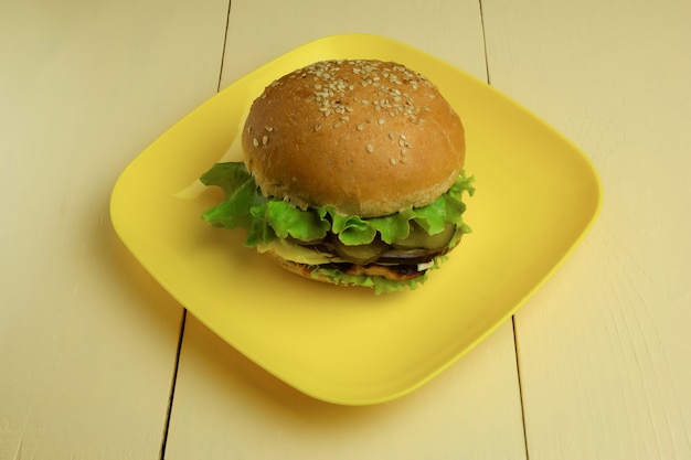 On a yellow ceramic square plate lies a large sandwich. A plate stands on a beige wooden table.