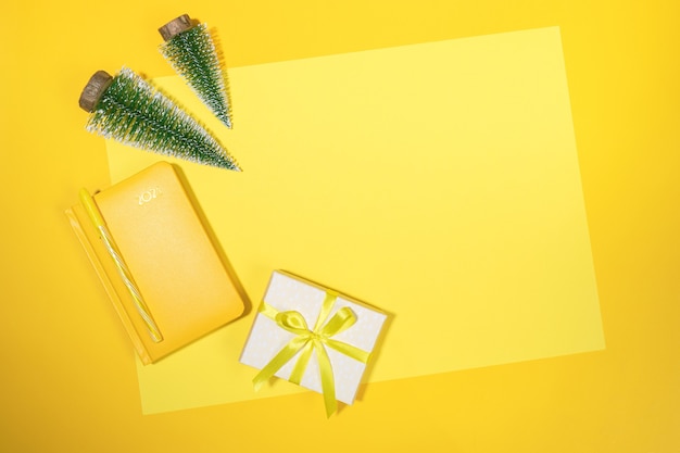 Yellow Christmas workspace backdrop. Frame of yellow notepad - organizer on 2021, small Xmas trees and DIY gift box on yellow sheet with copy space. Summing up, planning. Top view, flat lay.