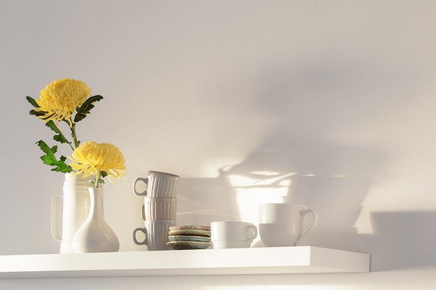 Yellow chrysanthemum in vase and cups on white shelf on white background