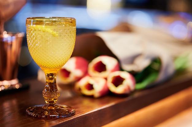 Yellow cocktail with lemon and ice in a beautiful glass medieval glass next to a bouquet of yellow-red Tulips