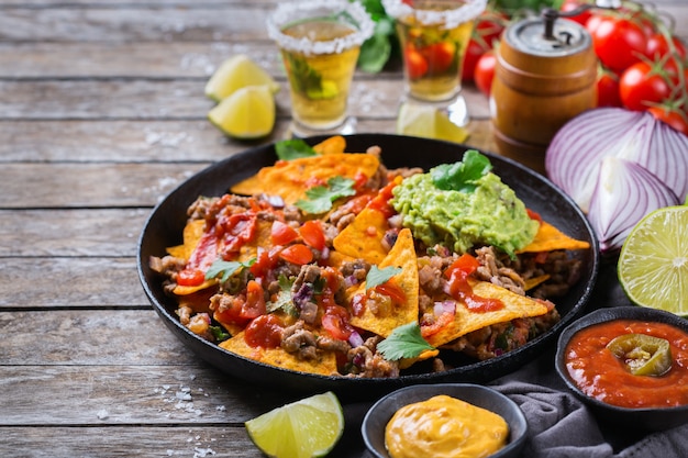 Photo yellow corn tortilla chips nachos with ground beef, mince, guacamole, red hot jalapeno chili salsa and cheese sauce with tequila on a wooden table.