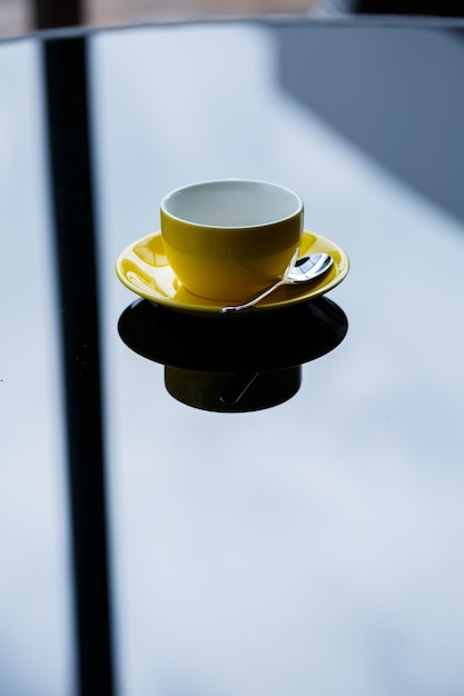 Yellow cup for coffee or tea with a saucer on a glass table.