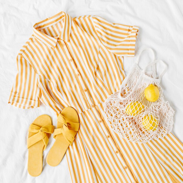 Yellow dress with stripes with eco bag and slippers on white bed. Women's stylish  summer outfit. Trendy clothes. Flat lay, top view.