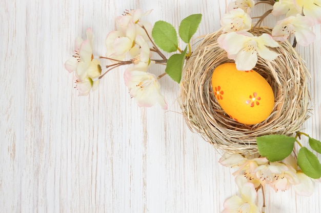 Yellow easter egg in straw nest and branch with flowers on white wooden background. top view, flat lay with space for text.