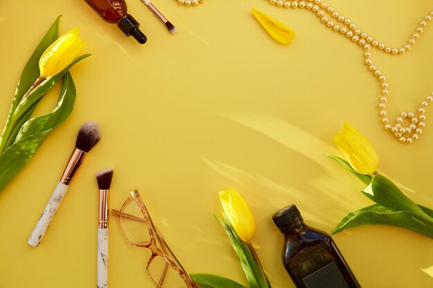 Yellow feminine flat lay makeup brushes glasses oil serum dropper pearls Yellow tulips and shadows Beauty card concept Copy space