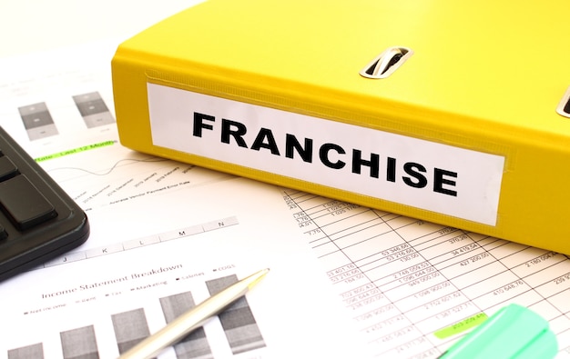 FRANCHISEというラベルの付いたドキュメントが入った黄色のフォルダは、財務チャートのあるオフィスの机の上にあります。