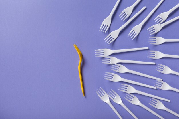 Yellow fork against lots of white plasic ones on purple background with copy space, top view. Opposition, be brave and different, leadership concept