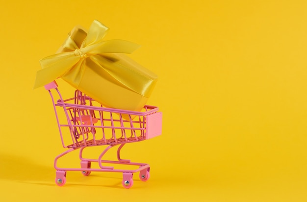 Yellow gift box with a silk bow in a miniature metal trolley on a yellow surface. party backdrop, surprise, seasonal sale