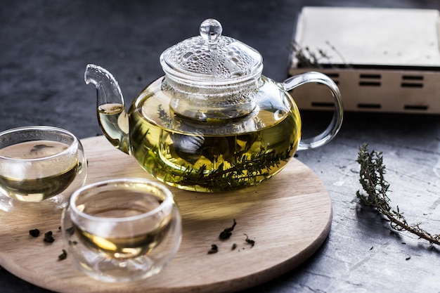 Yellow herbal tea in a transparent glass teapot and two bowls on a round wooden tray.