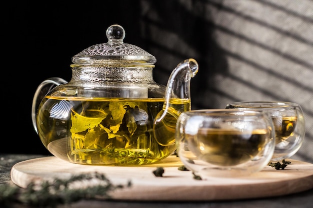 Yellow herbal tea in a transparent glass teapot and two bowls on a round wooden tray.